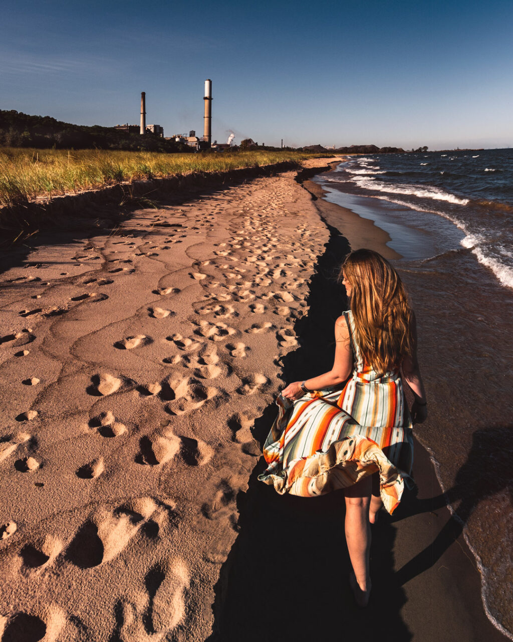 A woman in a colorful dress walking along the beach with a grassy plain, trees and an industrial harbor in the background.